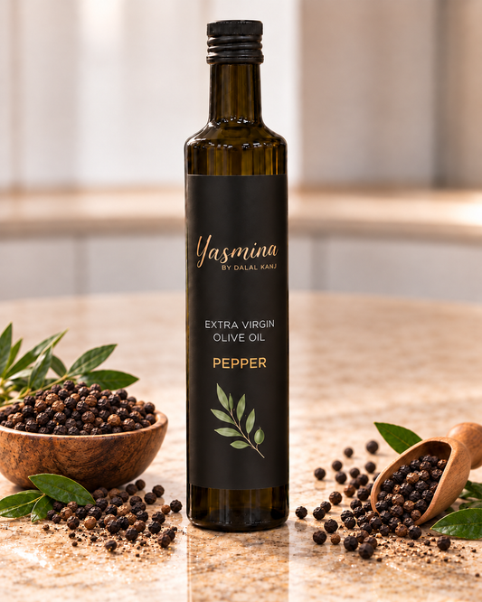A bottle of Yasmina by Dalal Kavi Extra Virgin Olive Oil Pepper, set on a marble countertop with a wooden bowl of peppercorns, scattered peppercorns, and green leaves.